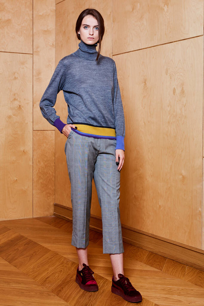 Studio portrait of a full-length stylish young girl in a gray trousers and a gray sweater on a plywood background - Foto, afbeelding