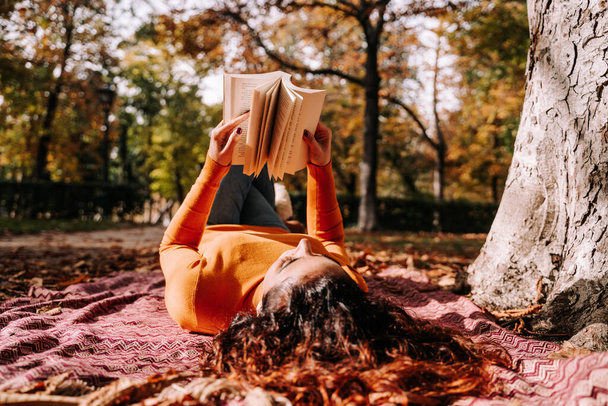 A pretty woman lying down on a blanket on the ground while reading a book. She is enjoying a break in a sunny autumnal day. Lifestyle autumnal outdoors - Photo, Image