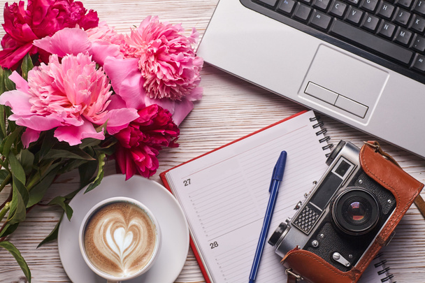Flach lag der Schreibtisch für Frauen. Weiblicher Arbeitsplatz mit Laptop, rosa Pfingstrosenstrauß, Kamera und Kaffee auf weißem Hintergrund. Weiblicher Hintergrund von oben. - Foto, Bild