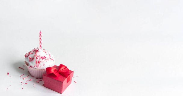 on a white background there is a cupcake with cream and 1 red candle and next to a small red gift with a bow  - Foto, Imagen