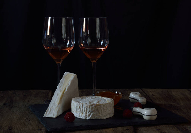 Black stone plate with french cheeses ,apicot marmalade,fresh raspberries, cheese  fork and cheese knife  and two glasses of  rose wine on rustic wooden table  - Photo, Image