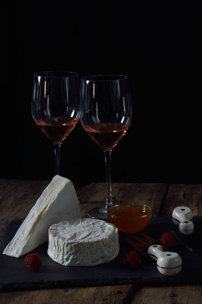 Černý kamenný talíř s francouzskými sýry, marmeládou, čerstvými malinami, sýrovou vidličkou a sýrovým nožem a dvěma sklenicemi růžového vína na rustikálním dřevěném stole  - Fotografie, Obrázek