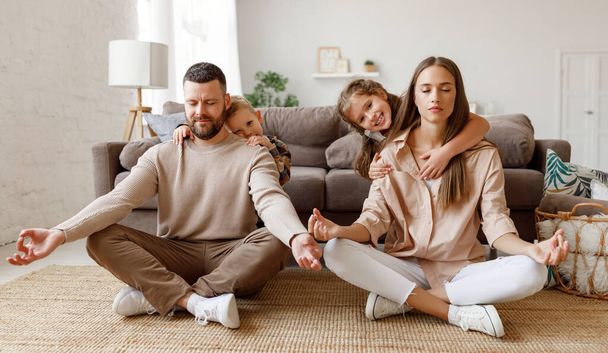 幸せな家族:静かな若い両親が蓮の位置に床に座って瞑想している間、子供たちは家で楽しさと抱擁しています - 写真・画像