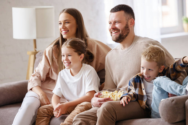 Rozkoszni rodzice i dzieci jedzą popcorn i śmieją się siedząc na kanapie i oglądając zabawny film w Hom - Zdjęcie, obraz