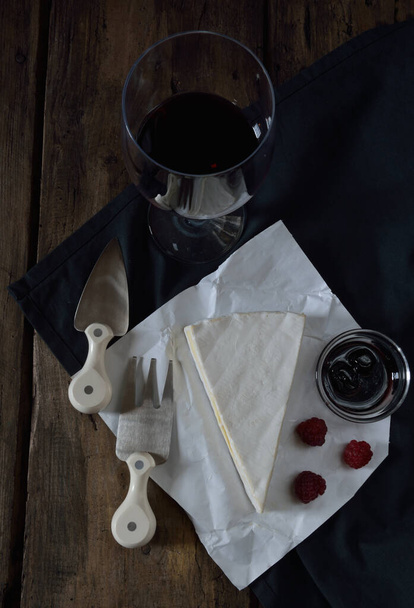 Brie-Käse, ein Glas Rotwein, Kirschlöffel süß, frische Himbeeren, Käsegabel und Käsemesser auf Holztisch - Foto, Bild