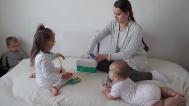 medicine, family, game concepts - Concentrated playful little kids wear medical glasses use stethoscope. Pretend be doctor, nurse, listen smile mommy.newborn baby sister breath deeply,sit on bed - Footage, Video