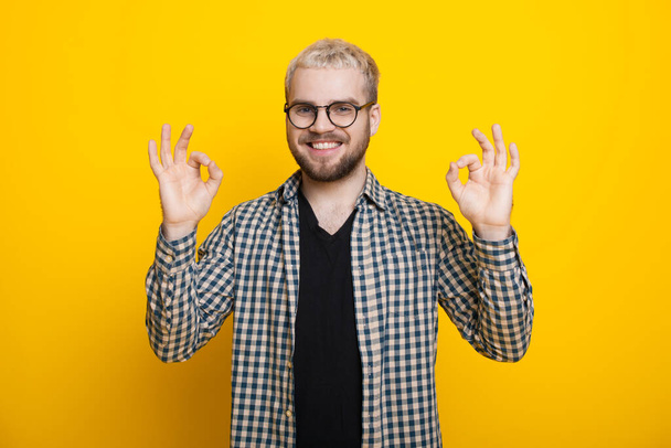 Biondo uomo con gli occhiali e la barba sta gesticolando il segno ok sorridendo alla fotocamera su un muro di studio giallo - Foto, immagini