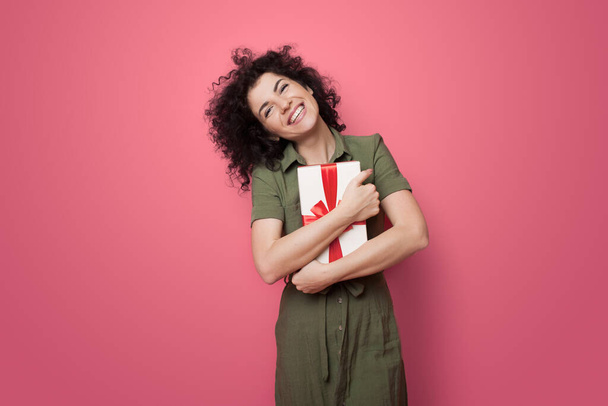 Улыбающаяся кудрявая женщина обнимает подарок на красной стене студии - Фото, изображение