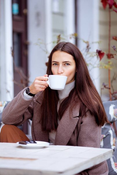 hosszú hajú barna nő kávét iszik egy utcai kávézó asztalánál. - Fotó, kép