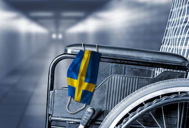 Flag of Sweden on face mask hanging on empty wheelchair in hallway of hospital or retirement nursing care home with copy space. Concept of COVID-19 pandemic in affecting seniors or elderly. - Photo, Image