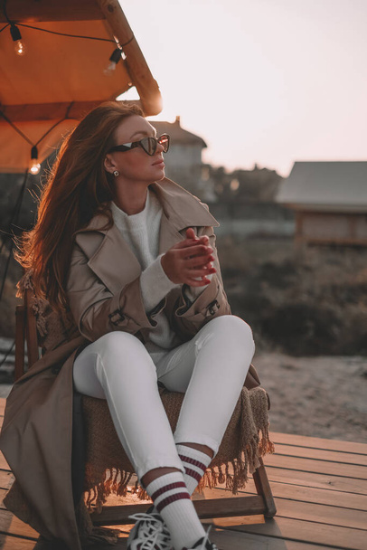 hermosa mujer se sienta en la terraza en un abrigo con gafas mira a la puesta de sol. chica descansando en una casa de campo sentado en una silla de madera - Foto, imagen
