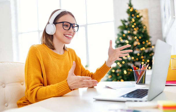 Cheerful woman in glasses and headphones smiling and speaking with online colleague on Christmas day at hom - Foto, imagen