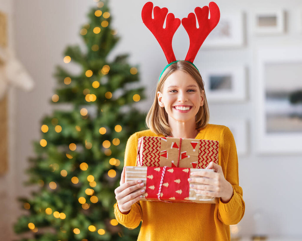 szczęśliwa młoda kobieta z pakowane prezenty na święta Bożego Narodzenia uśmiecha się do kamery w hom - Zdjęcie, obraz