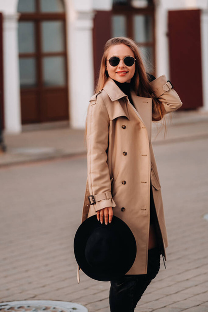 Stylish young woman in a beige coat and a black hat in his hands and glasses on a city street. Women's street fashion. Autumn clothing.Urban style - Photo, Image