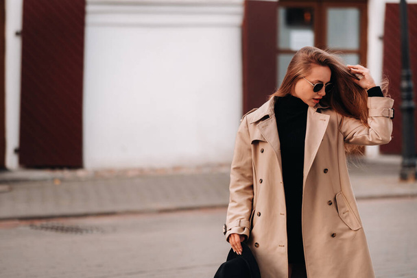 Stylish young woman in a beige coat and a black hat in his hands and glasses on a city street. Women's street fashion. Autumn clothing.Urban style - Valokuva, kuva