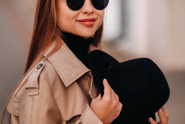 Stylish young woman in a beige coat and a black hat in his hands and glasses on a city street. Women's street fashion. Autumn clothing.Urban style - Foto, Imagem