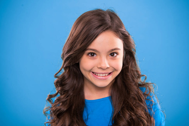 Perfect curling hair. Kid girl long healthy shiny hair. Kid happy cute face with adorable curly hairstyle stand over blue background. Teaching healthy hair care habits. Little girl grow long hair - Valokuva, kuva