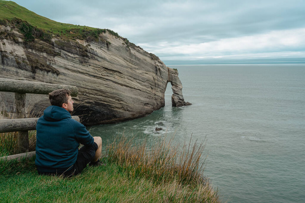 Νεαρός Καυκάσιος κάθεται στην κορυφή του γκρεμού και κοιτάζει το ακρωτήριο Αποχαιρετισμός σχηματισμού βράχου. Νέα Ζηλανδία. - Φωτογραφία, εικόνα