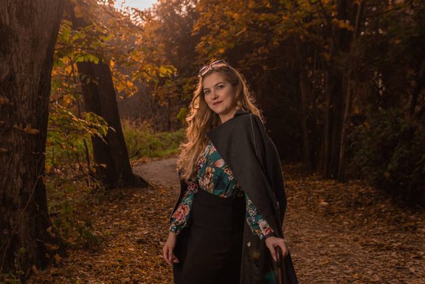 A beautiful young girl in a black cloak stands in glasses chameleons in the fall. Yellow forest, green grass, blonde. smiling at camera urban life concept - Фото, зображення
