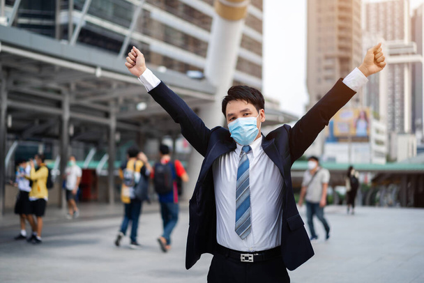 succesvolle zakenman met opgeheven armen en het dragen van medisch masker tijdens de coronavirus (covid-19) pandemie in de stad - Foto, afbeelding