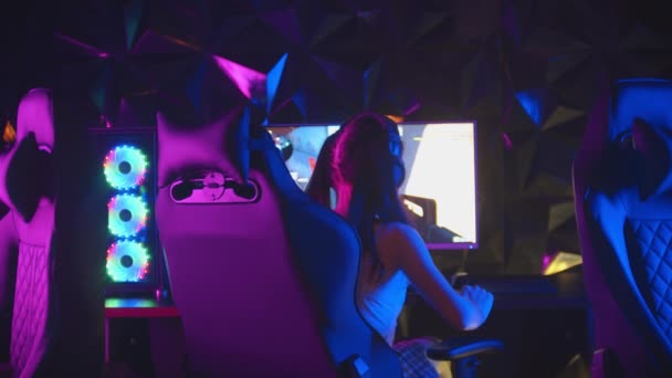 Junge hübsche Frau sitzt im neonfarbenen Spielclub und spielt Spiele - dreht sich vom Monitor ab, nimmt ihre Kopfhörer ab und blickt in die Kamera - Filmmaterial, Video