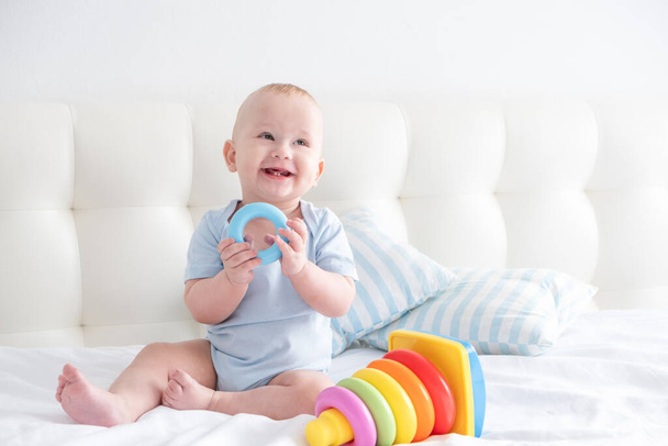 grappig baby jongen zit op bed en spelen met een speelgoed kinderen piramide. - Foto, afbeelding