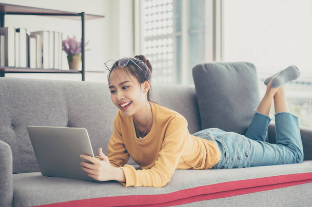 Beautiful young asian woman lying down and using computer laptop talking video conference call on sofa in living room - Foto, Imagem