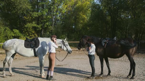 Due cavalleresche in abiti da equitazione e caschi si preparano a cavalcare sui loro cavalli - Filmati, video