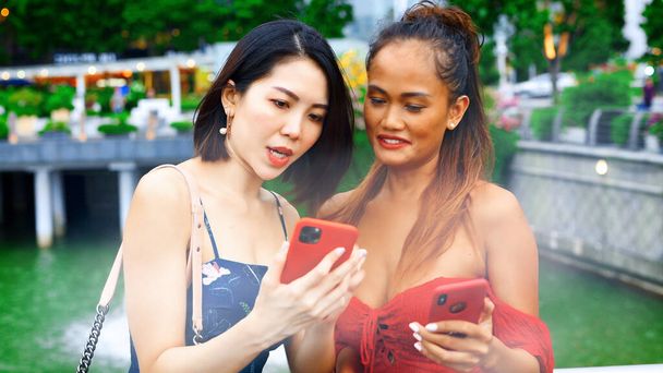 2人の美しい若いアジアの女性が話して、笑顔で、街の通りを歩いてリラックスし、彼らのスマートフォンを見て. - 写真・画像