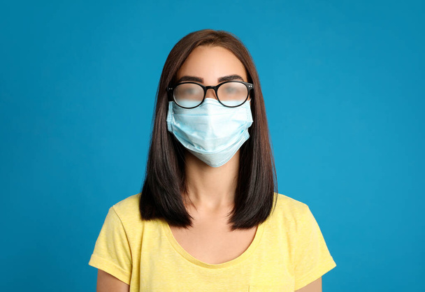 Young woman with foggy glasses caused by wearing disposable mask on blue background. Protective measure during coronavirus pandemic - Valokuva, kuva