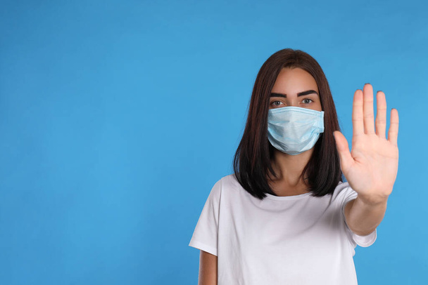 Young woman in protective mask showing stop gesture on light blue background, space for text. Prevent spreading of coronavirus - Foto, immagini