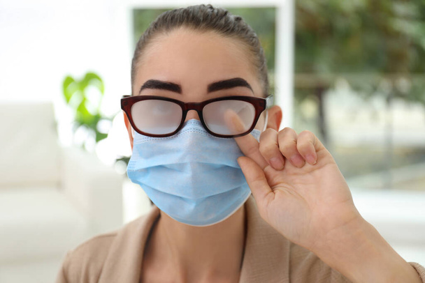 Woman wiping foggy glasses caused by wearing medical mask indoors, closeup - Foto, Imagen