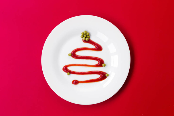 Ketchup in a white plate looks like a Christmas tree. Green peas serve as a decoration. The plate stands on a red background. View from above. Horizontal photo - 写真・画像