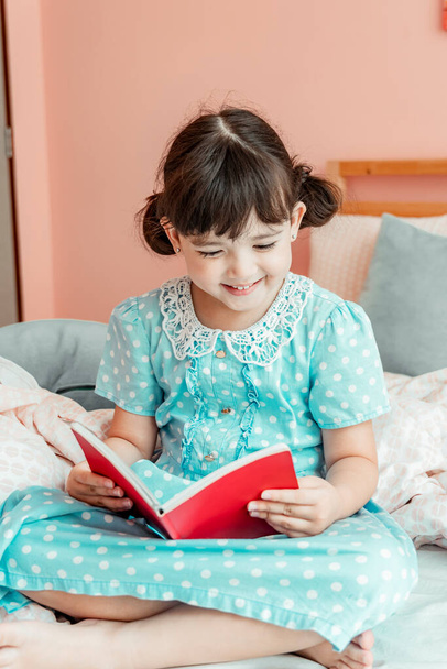 Fille heureuse en robe bleue lisant un livre dans la chambre. - Photo, image
