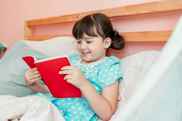 Adorable chica en vestido azul leer un libro antes de acostarse en el dormitorio. - Foto, Imagen