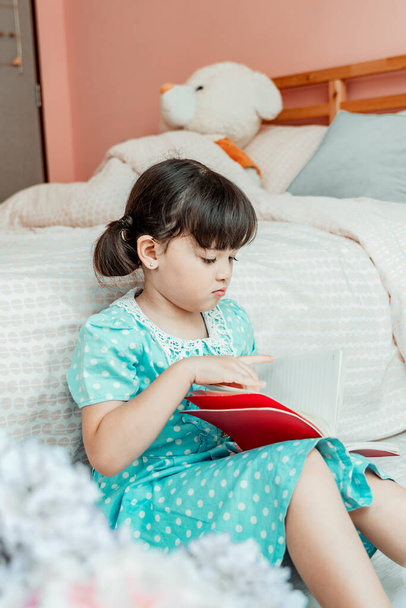Söpö pieni tyttö sininen mekko lukeminen kirja makuuhuoneessa. - Valokuva, kuva