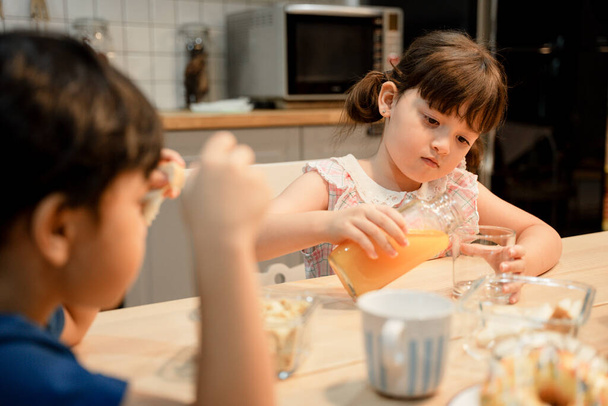 Hermana vertiendo jugo de naranja para servir a su hermano en la cocina. - Foto, imagen