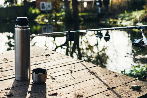 A thermos with hot tea, coffee is on a wooden deck on the shore of the lake and next to a fishing rod. Travel alone, outdoor recreation, halt, rest, fishing. - Photo, Image