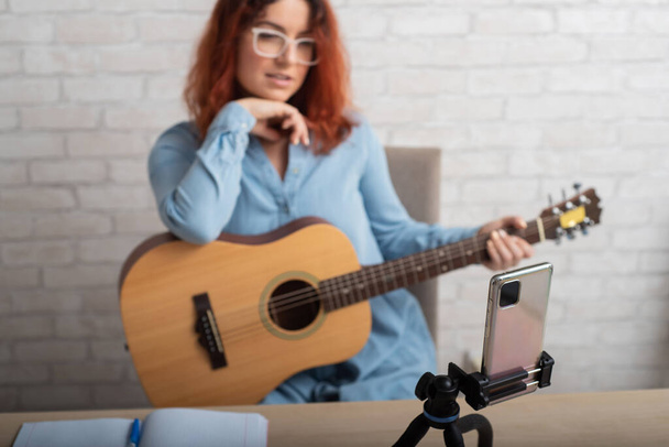 An attractive red-haired woman blogs on music. The girl sings, plays the guitar and records video on a smartphone - Φωτογραφία, εικόνα