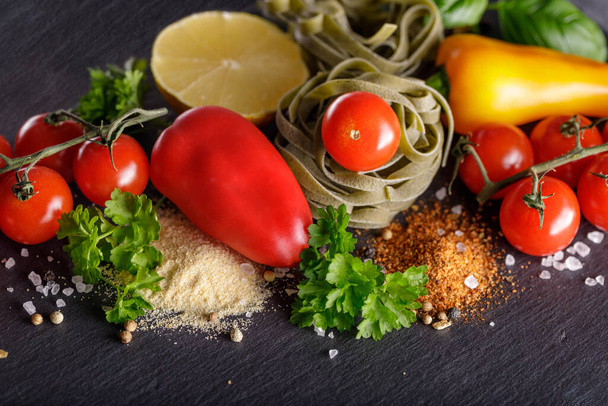 Cherry tomatoes, multicolored peppers, pasta spinach tagliatelle, parsley, slice of lemon and spices on a stone board. Dark background with copy space - Фото, изображение