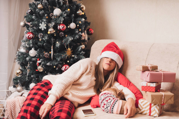Woman falling asleep and tired of gift wrapping with puppy dog on a couch in the living room with Christmas tree at home - Foto, afbeelding