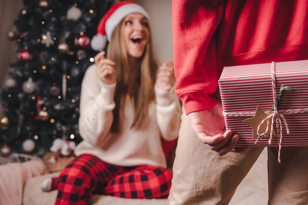 Şaşırtıcı bir şekilde kırmızı kazaklı bir adamın Noel hediyesi kutusunu arkasındayken süslenmiş Noel ağacının yanında dikiz aynasından bakması. - Fotoğraf, Görsel