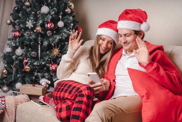 Portrait of lovely couple husband and wife in red Santa hat having a video call or taking selfie with their smartphone, enjoy Christmas time at home - Photo, image