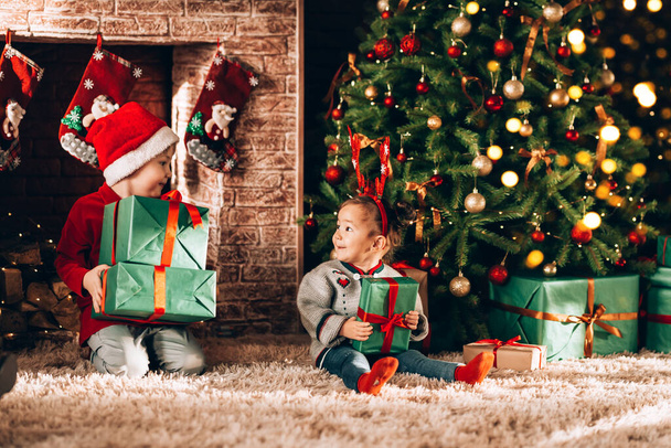 Bruder und Schwester sitzen mit Kartons in grüner Verpackung mit Geschenken und einer Verbeugung. Weihnachtsstimmung. Kinder in Neujahrskostümen warten auf ein Wunder. Vor dem Hintergrund von Weihnachtsbaum und Kamin. - Foto, Bild