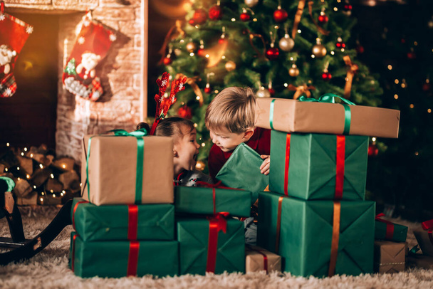 Brother and sister are sitting with boxes in green packaging with gifts and a bow. Christmas mood. Children in New Year's costumes waiting for a miracle. Against the background of a Christmas tree and fireplace. - Photo, image