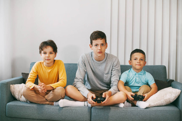 Three of the teen's best happy friends are playing a video game at home. Three brothers play a video game, sitting on the couch, during the Christmas holidays. Family holiday, time together. - Foto, Imagem