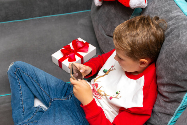 Κοντινό πλάνο Ελκυστικό παιδί αγόρι που χρησιμοποιεί smartphone για τις γιορτές των Χριστουγέννων λευκό δώρο για τη φωτογραφία x-mas στο σπίτι. Εικόνα πλευρικής προβολής. - Φωτογραφία, εικόνα