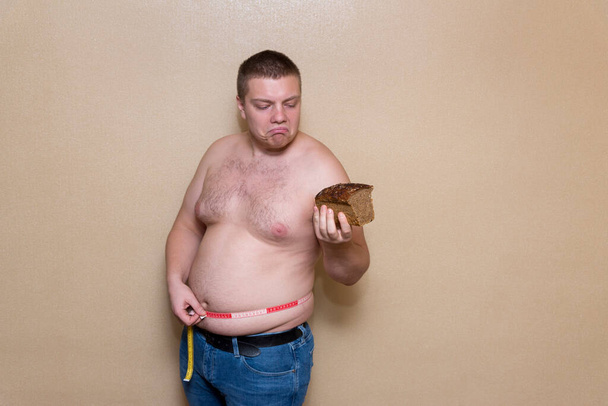 Nuori mies mittaa lihavan vatsansa mittarilla ja pitää leipää yhdessä kädessä. - Valokuva, kuva