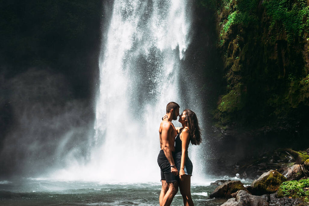 Beautiful couple kissing at the waterfall. A couple in love at a beautiful waterfall. Wedding trip to Bali. Honeymoon trip. A man of athletic build kisses a beautiful woman. Vacation in Bali. - Фото, зображення