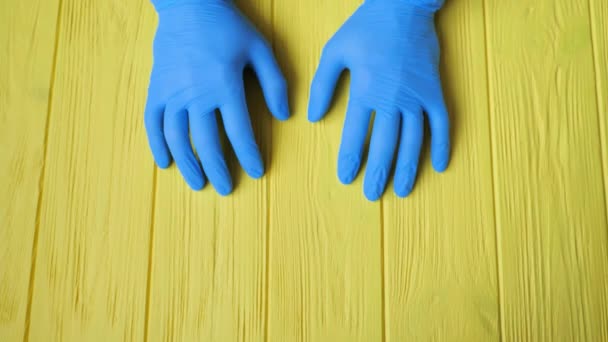 Manos de guante azul cerca de la mesa de madera amarilla. Pide desinfección de manos. Detener el concepto de coronavirus. Uso de gel antibacteriano para las manos durante la cuarentena - Metraje, vídeo